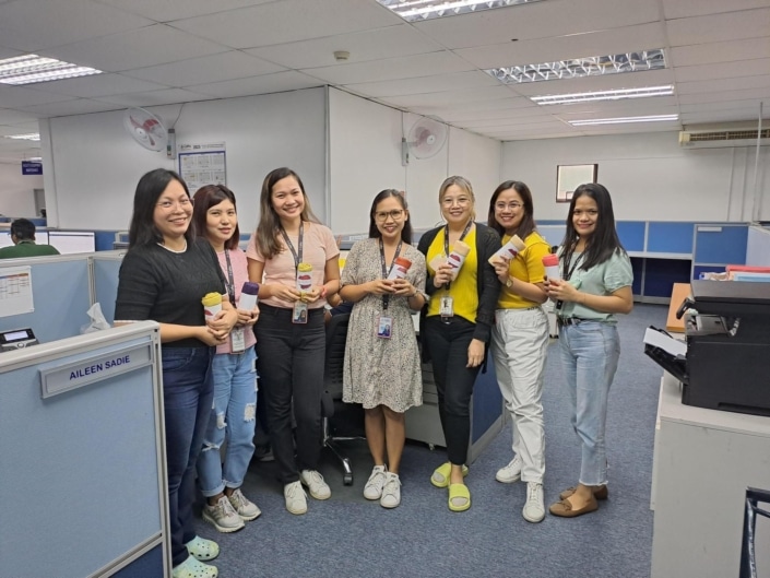 A group of seven women smiling and each holding a rolled up shirt. They are in an office and standing outside of a cubicle.