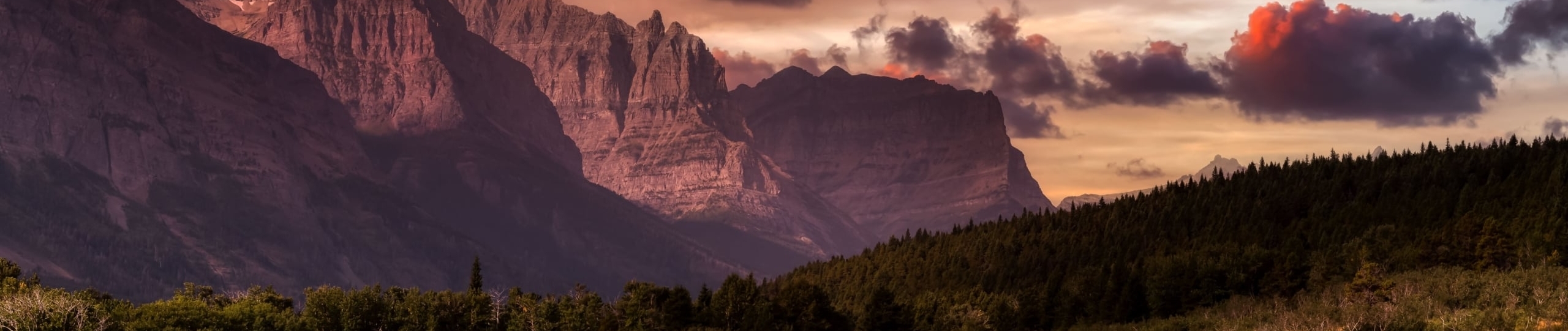 A rustic landscape just before sunset of towering rock mountains and lush green forests in the foreground.