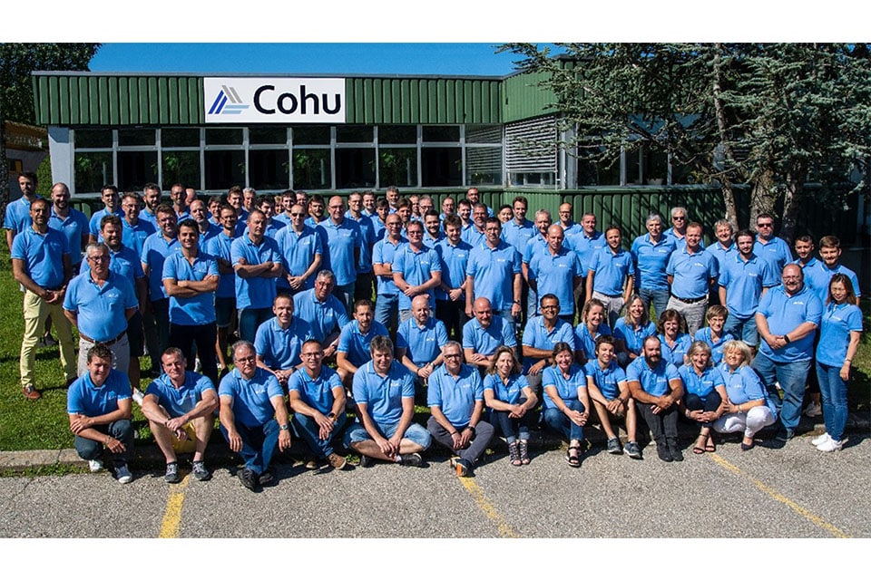 A group of approximately 80 people wearing light blue shirts smiling outside in front of a building that has a sign that reads Cohu.