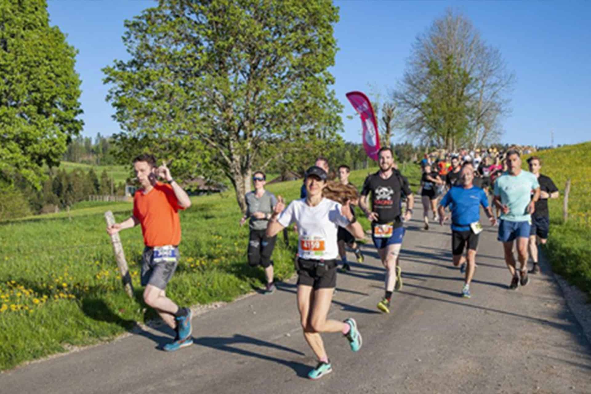 A group of over 20 people running in a race along a paved road surrounded by greenery and race flags lining the path.