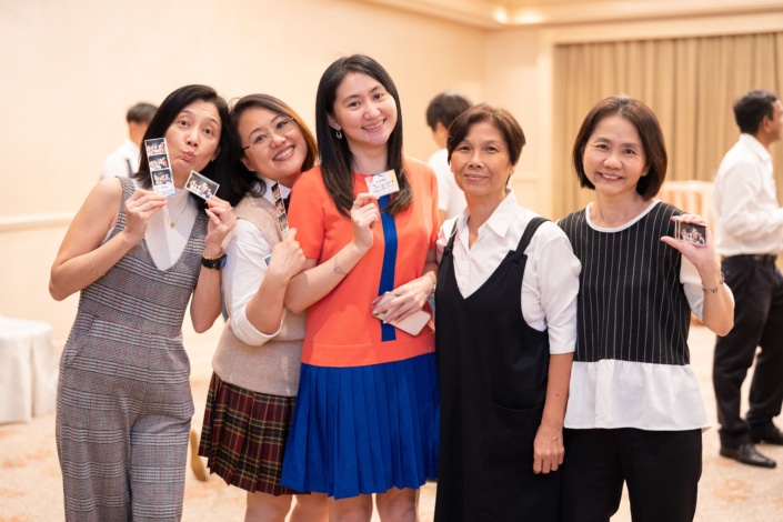 A group of five women dressed formally and smiling. They are in a banquet hall and are holding up photographs.