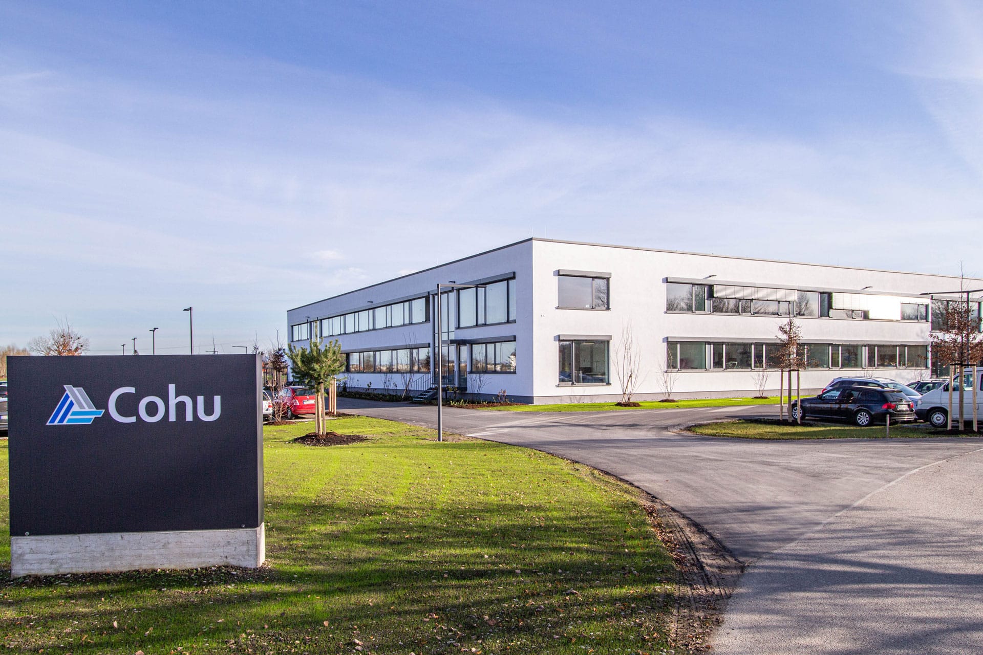 A two story white building lined with windows set in the background. A black sign that reads Cohu and a green grass lawn are in the foreground with cars off to the right.