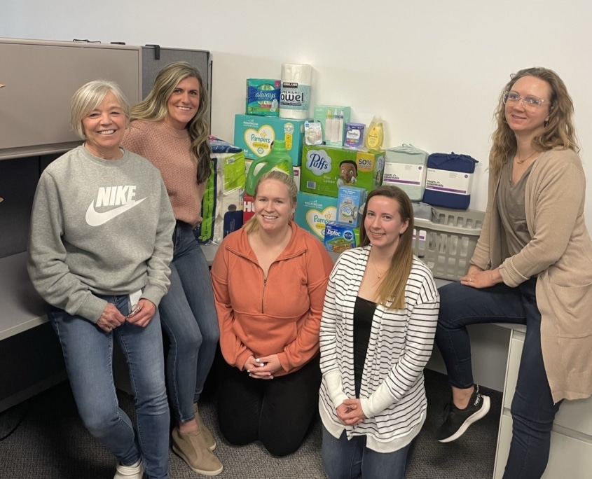 A group of 5 women smiling and standing in front of a pile of tissues, diapers, sheets, soap, paper towels. They are indoors in an office setting.