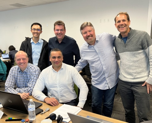 A group of 6 men standing around an office table that has laptops, papers, and other office supplies on it. They are smiling and two are sitting at the table.