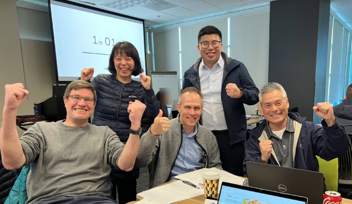 A group of five people smiling and holding their fists up in excitement. Three are sitting at a table and two are standing and there is a projector screen behind them that reads 1m 01.