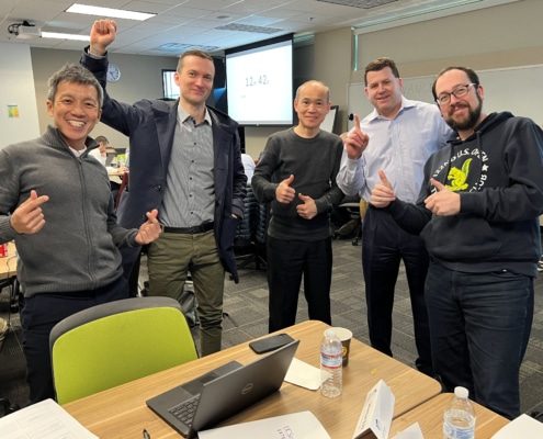 A group of 5 men smiling in front of an office table and giving thumbs ups. There are groups of others at desks in the background working.