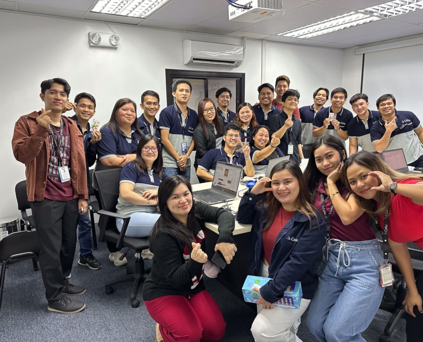 A group of 22 people smiling and holding peace signs and making hearts with their fingers. They are in an office environment, some are standing behind an office table and some are in front of it.