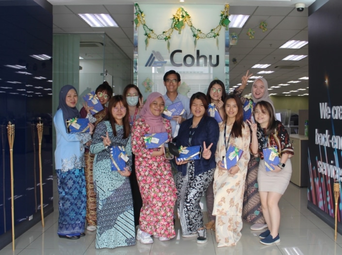 A group of eleven people standing together indoors and showing peace signs. They are all holding blue boxes with a gold ribbon on them.