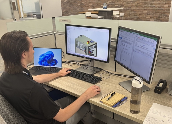 A man sitting at a desk with three monitors in front of him, each displaying a different screen. He is in an office environment with a water bottle and notebook to his right.