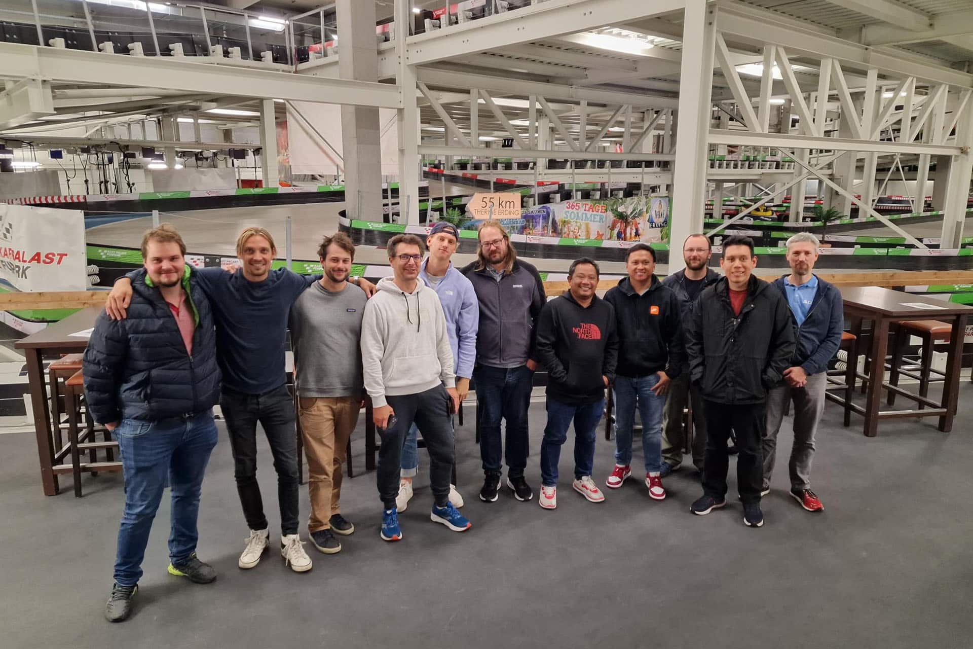 A group of 11 men standing together smiling. They are standing indoors in front of an indoor race track with tables and chairs beside them.