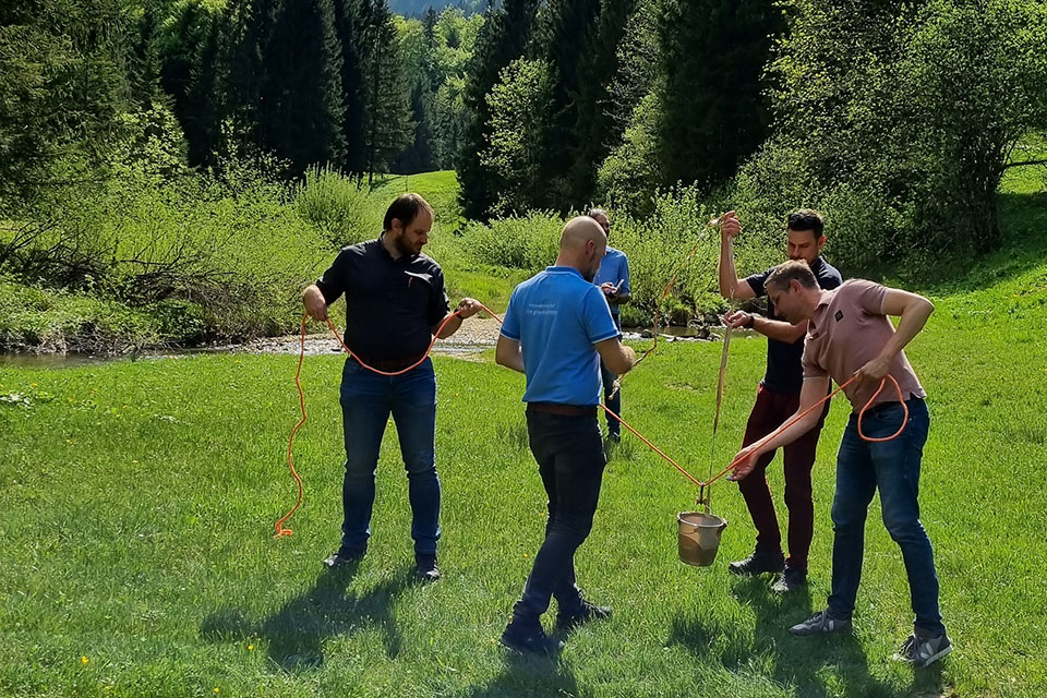 A group of 5 men standing together in an open field with trees in the background. Three of the men are holding ropes that are attached to a bucket at the other end.