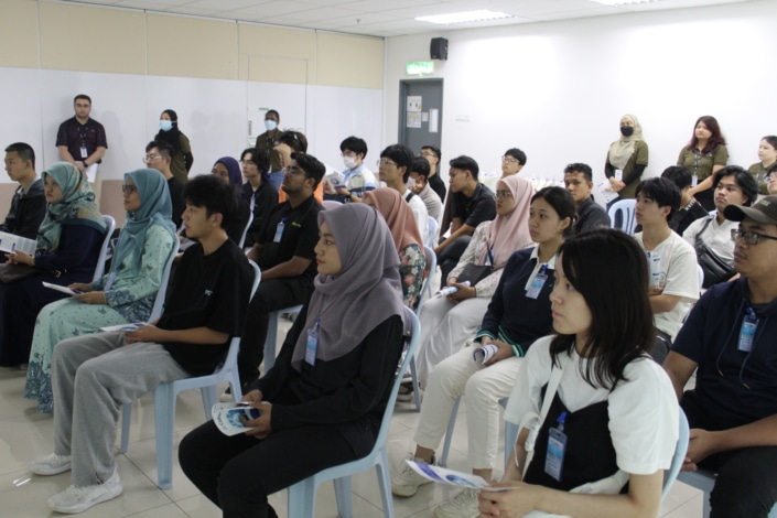 A group of 32 people are gathered in a room holding brochures and listening intently towards the front of the room. Six of the individuals are standing and are standing along the edges of the room.