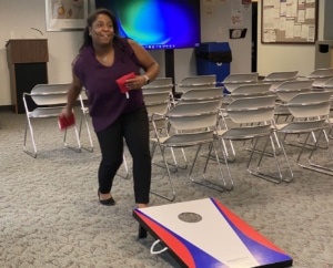 A woman playing cornhole indoors and about to toss a bag. Behind her, are three rows of chairs and a large TV screen.