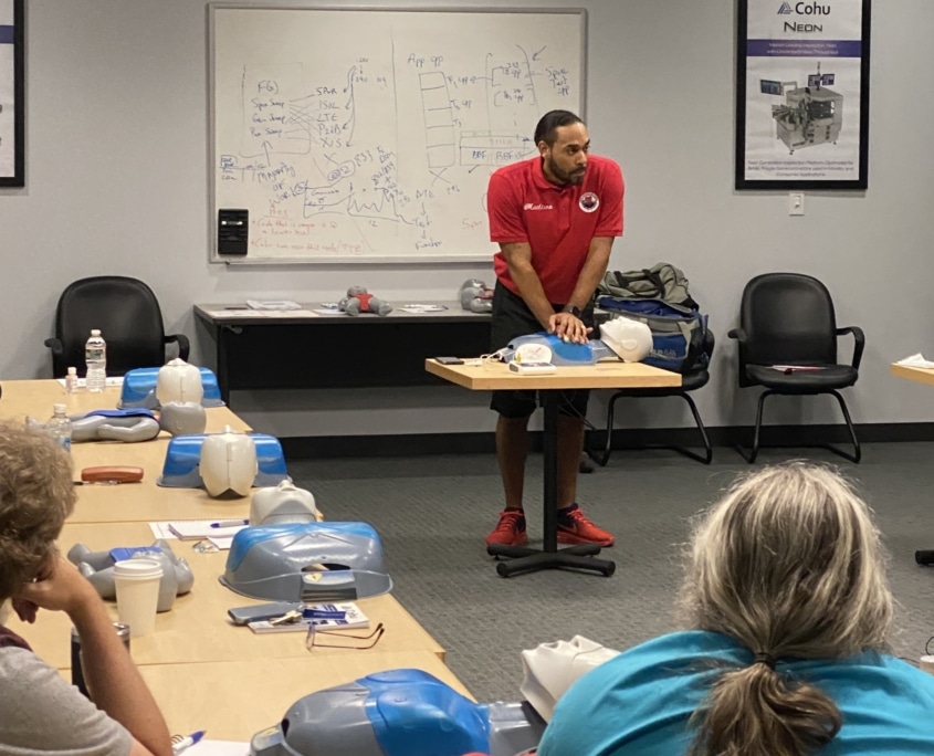 A man in a red shirt leaning on a plastic dummy of a chest and head used in CPR training. There is a long table to the man's left with four more dummies laid out for practicing.