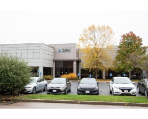 A one story beige building with a Cohu sign with trees on either side of the entrance and four cars in the foreground.