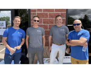 A group of 4 men standing, smiling in front of a brick building and glass windows. Two men are wearing grey shirts and two are wearing blue shirts.