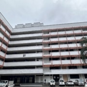 A long seven story building with red and white accents on a cloudy day. There are at least six cars in the parking lot in front of the building.