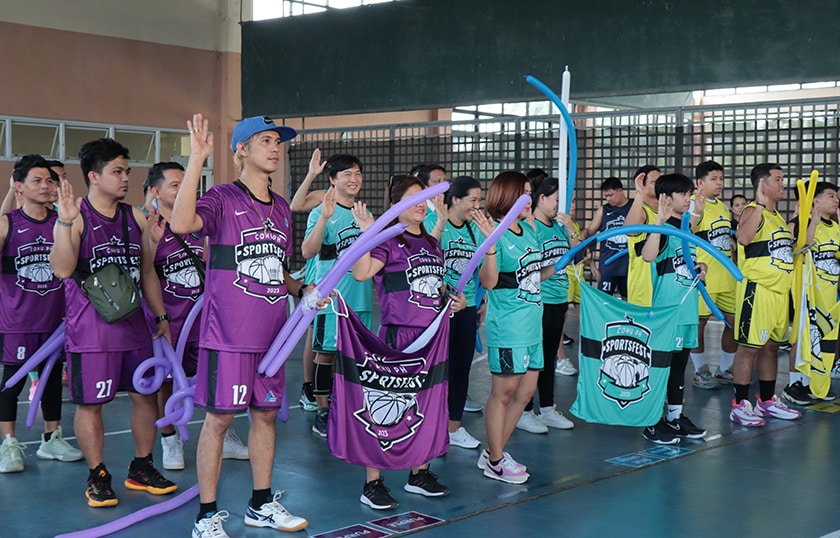 Approximately 21 people standing in a gymnasium holding up their right hands and holding balloons in their left hands. They are wearing coordinated colorful team jerseys and there are 3 different teams visible.