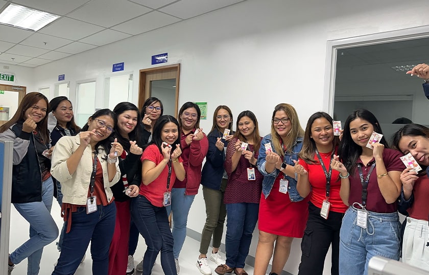 A group of 13 women standing in a line together, smiling and making heart signs with their fingers in an office environment.