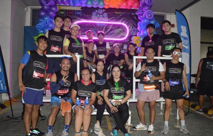 A group of 17 people standing together smiling wearing black shirts. They are surrounded by bright colored balloons and neon lights.