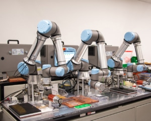 Three robotic arms hovering over a table. The table is filled with probes and various containers and wires lying around.