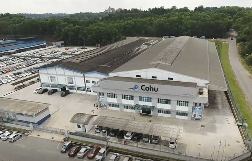 An aerial image of a long three story building with a full parking lot in the left side of the image. There is a road in the front of the building and to the left.