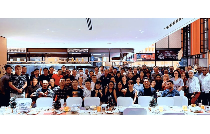 A group of about 58 people smiling and standing in front of a long table with a white tablecloth in a restaurant.