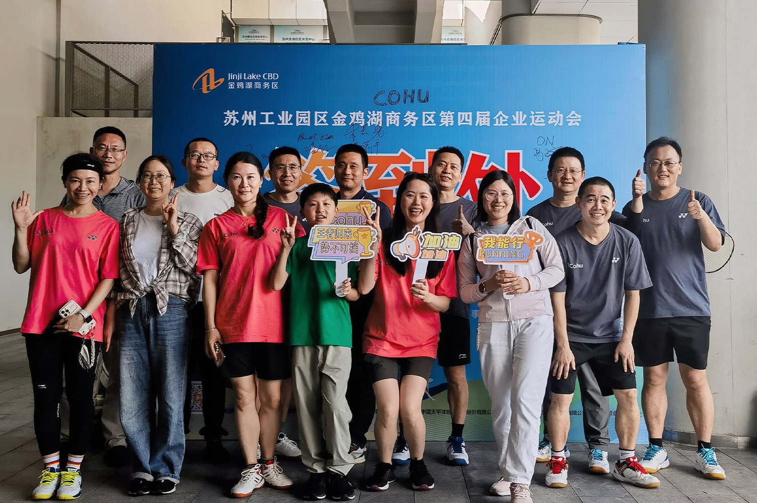 A group of 14 people smiling and four holding signs in Chinese. They are standing in front of a blue backdrop banner with Chinese characters on it.