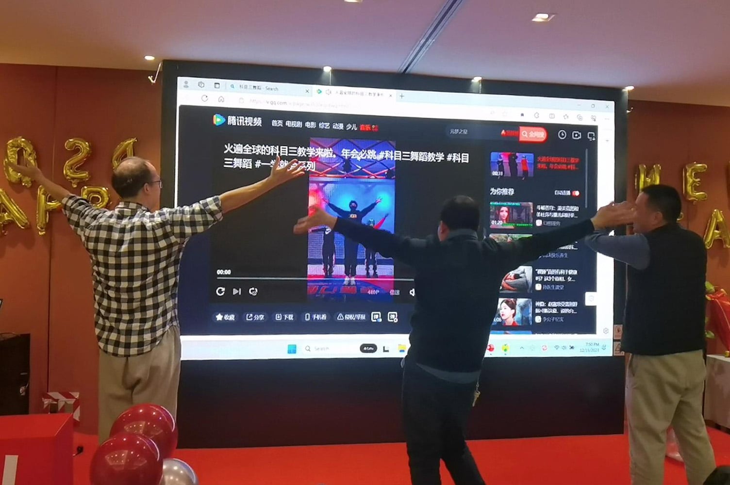 Three men standing in front of a projector screen with their arms outstretched. The projector is showing a dance video in Chinese. The carpet is red and there are balloons around the room.