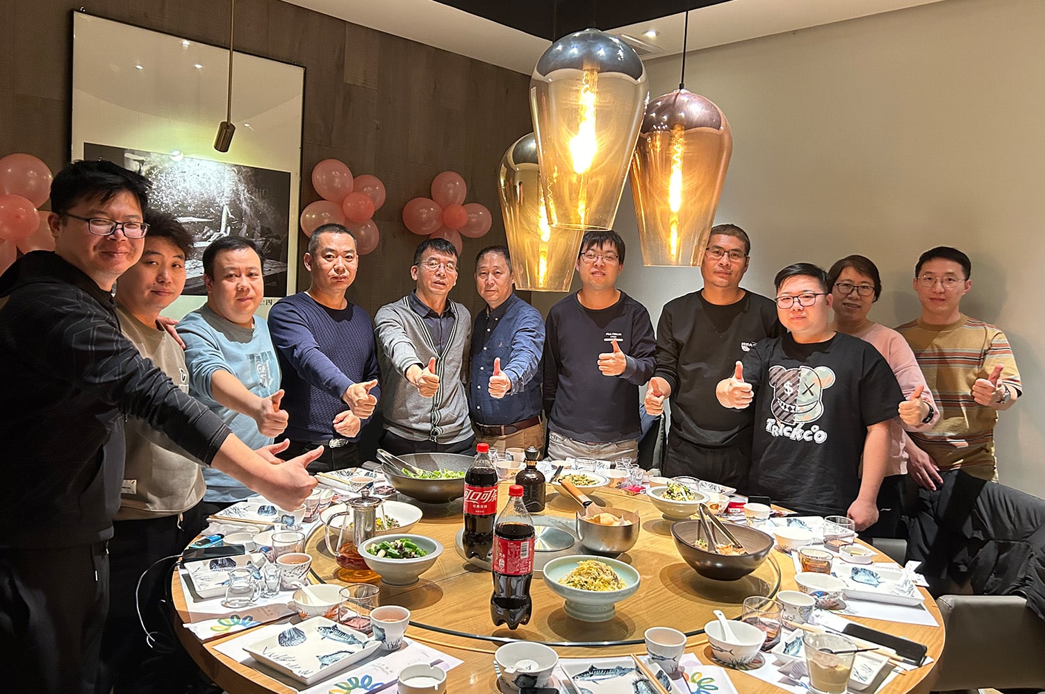 A group of eleven men standing around a round table with food and dishes. The men are all looking at the camera and giving thumbs ups.