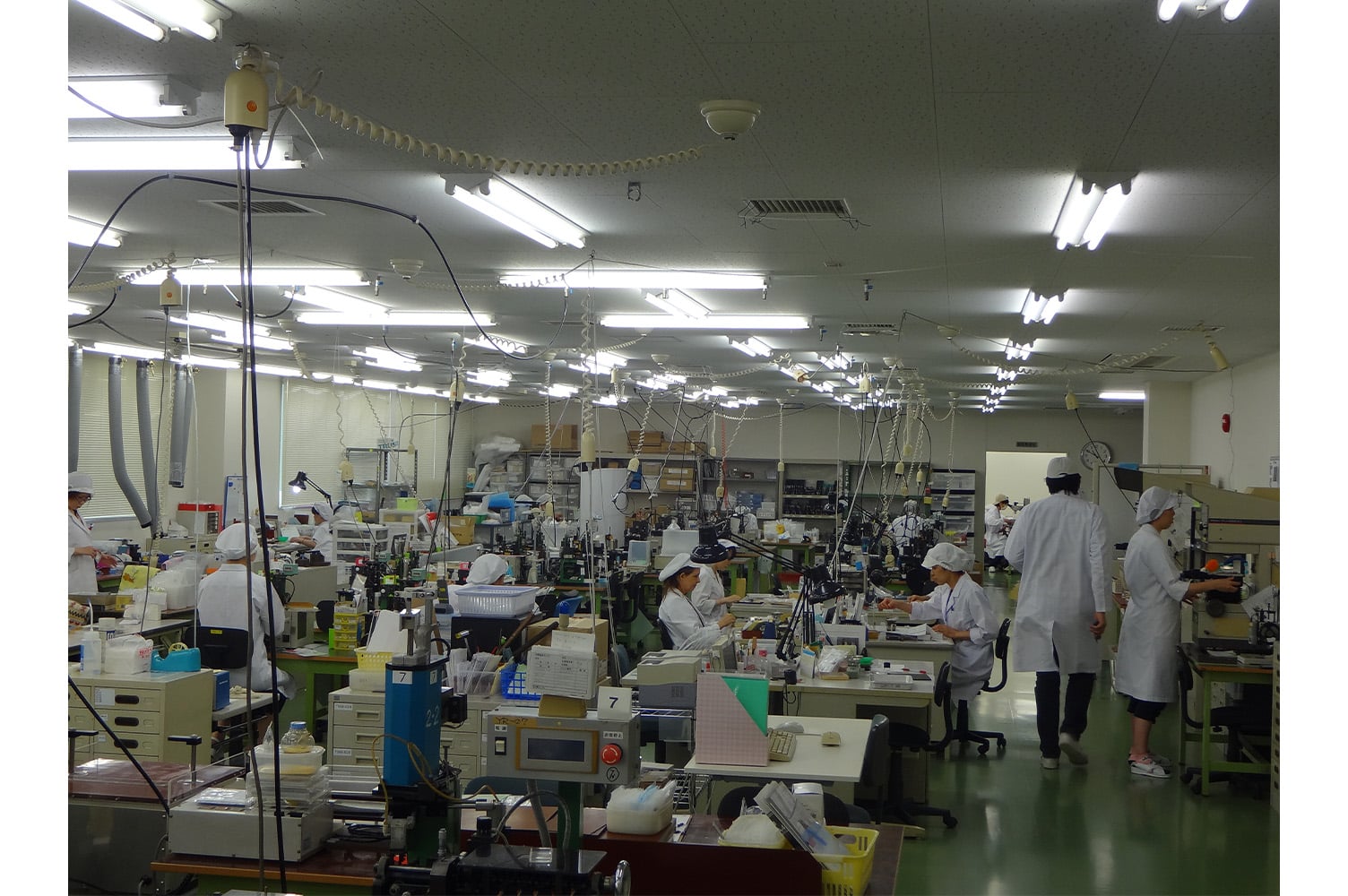 A cluttered production floor with people in white coats, sitting and standing, and cords hanging from the ceiling.