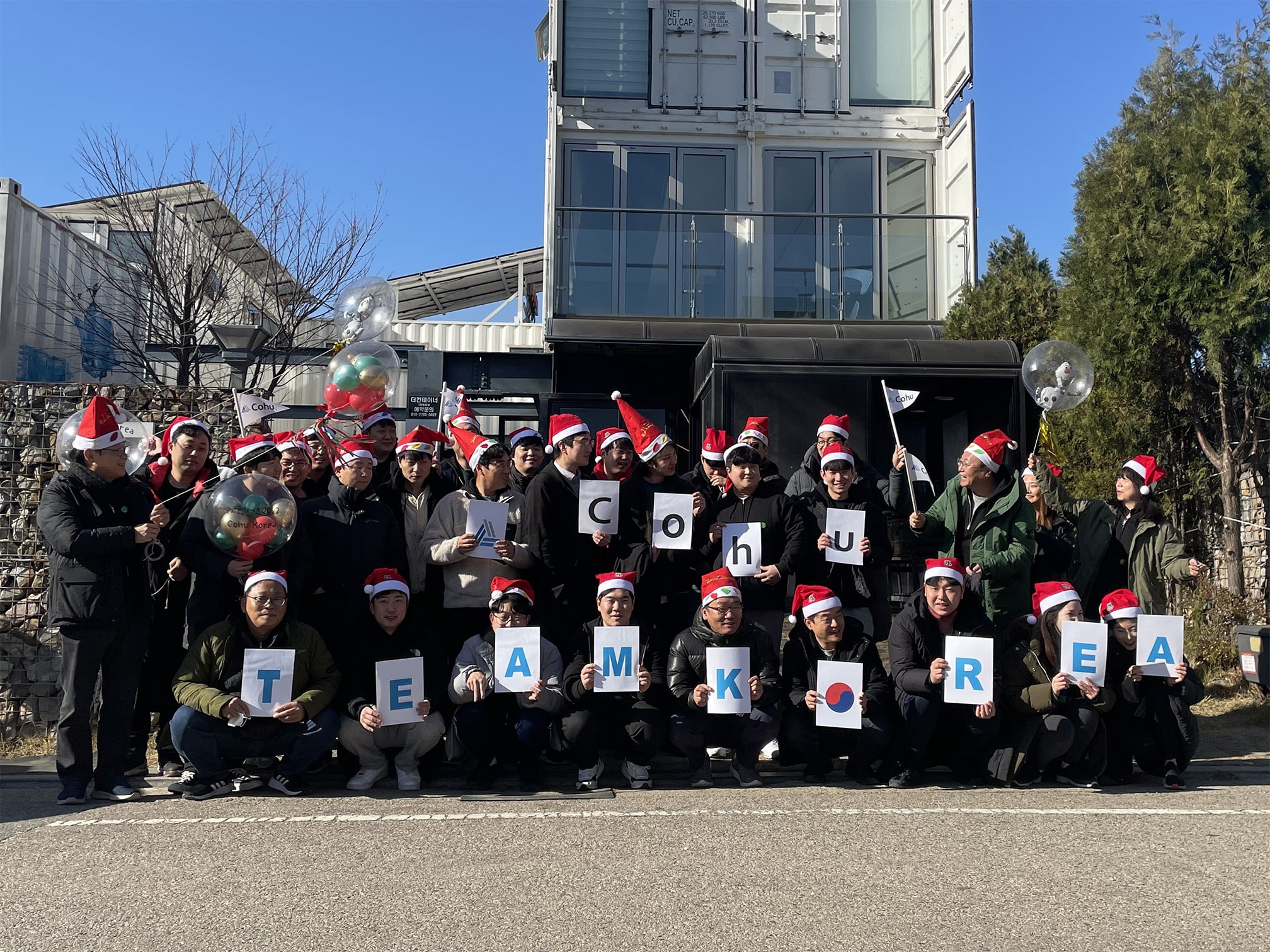 A group of 31 people smiling outside wearing santa hats and holding individual letters that spell out Cohu Team Korea.