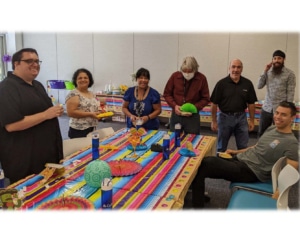 A group of seven people standing around a table with a multi-colored tablecloth. Six of them are smiling and four of them are holding decorations for the table.