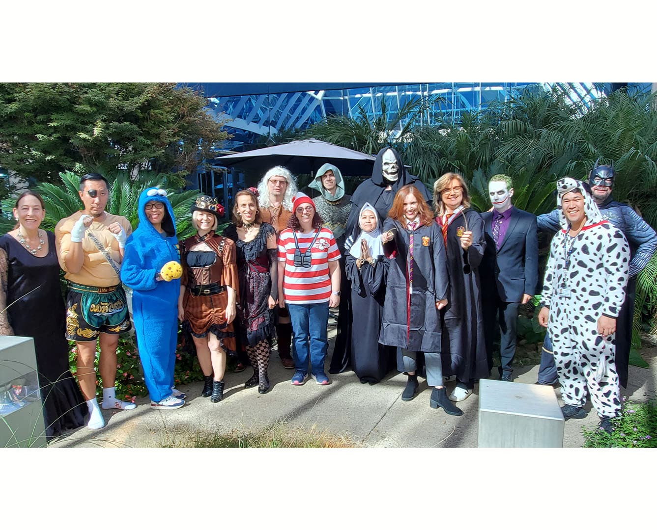 A group of 15 people smiling dressed in halloween costumes in a greenhouse setting with plants behind them.