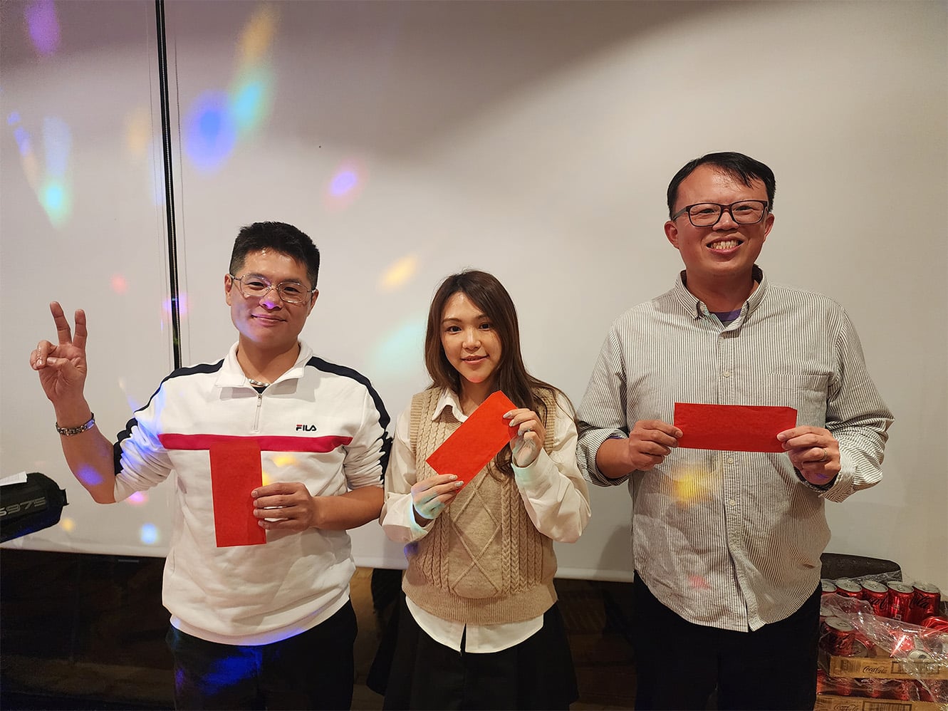 Two men and one woman smiling and holdin up red envelopes. One man is holding up the peace sign.